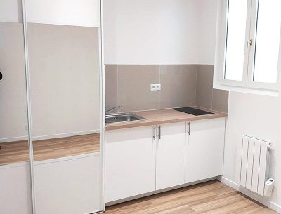 Vue de la pièce principale rénovée avec cuisine équipée, sol en carrelage imitation parquet et peinture neuve.