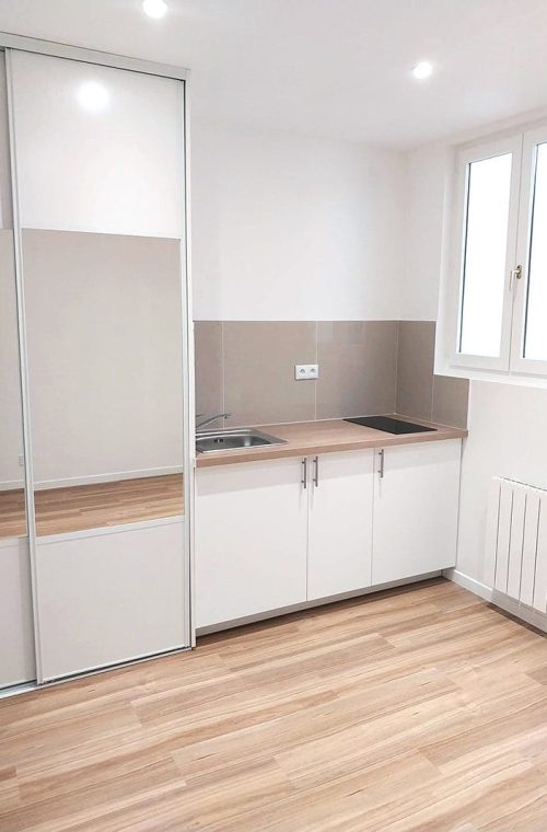 Vue de la pièce principale rénovée avec cuisine équipée, sol en carrelage imitation parquet et peinture neuve.